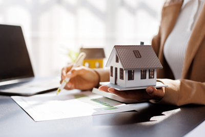 person holding model house while analyzing documents at desk with laptop displaying home investment growth trends